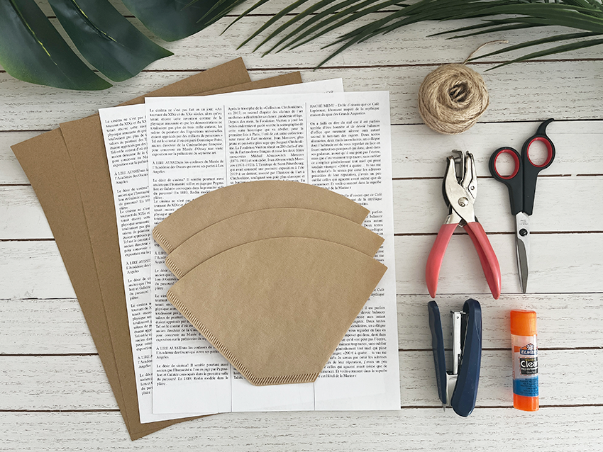 Tools and materials used to create small treat bags made with coffee filters, including coffee filters, jute cord, hole puncher, jute cord, glue stick, scissors, kraft paper and newspaper print